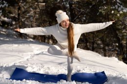 Девушка в белом свитере и шапке занимается йогой на коврике на улице зимой